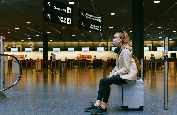 Ragazza con mascherina anti covid seduta su una valigia in aeroporto deserto